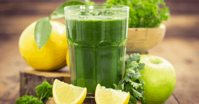 Green smoothie with fruit and greens on counter around the glass.