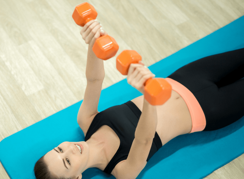 lady laying on yoga mat on floor doing bench press with two dumbbells