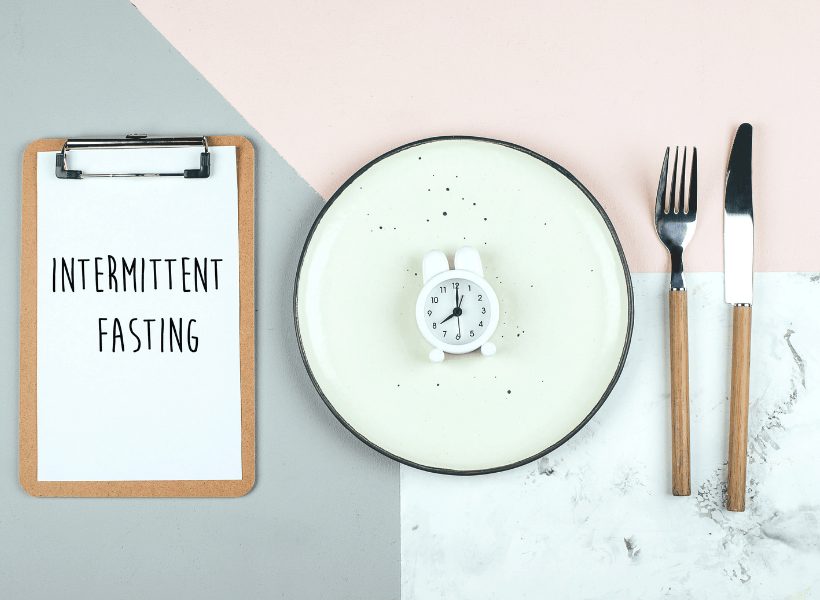 clipboard with white sheet that says intermittent fasting beside a green plate with a clock beside a fork and knife