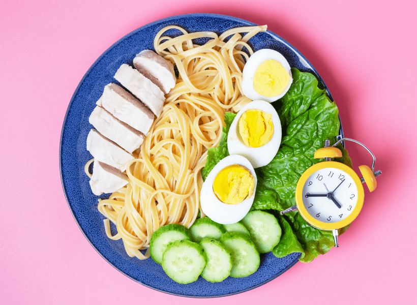 blue plate full of rows of food that include chicken, pasta, boiled eggs, cucumbers and lettuce with a small yellow and white clock on the corner