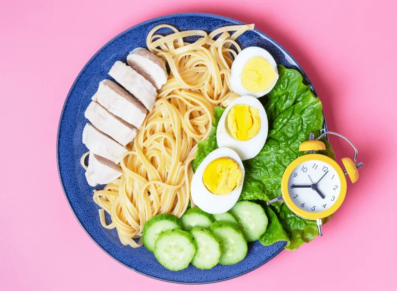 blue plate full of rows of food that include chicken, pasta, boiled eggs, cucumbers and lettuce with a small yellow and white clock on the corner