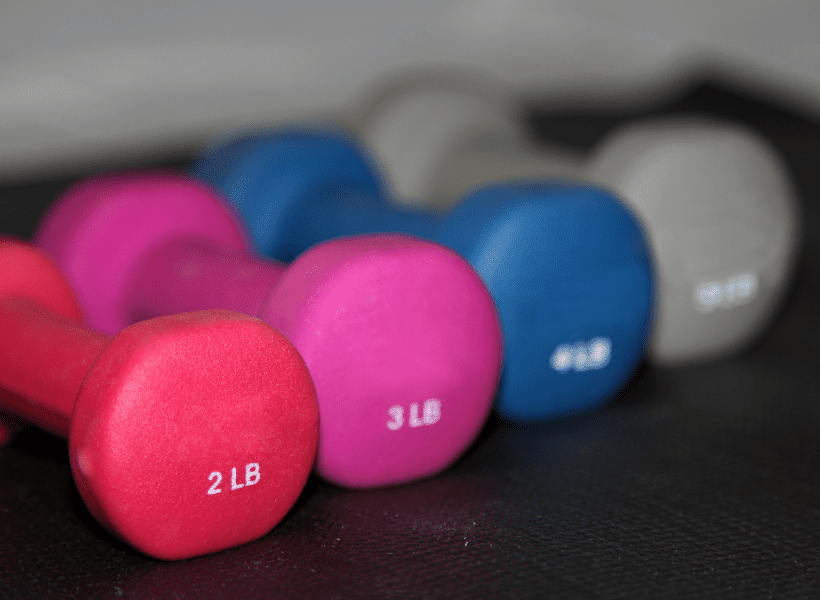 four single colored dumbbells 2 lb red hand weight, pick 3 lb hand weight, 4 lb blue hand weight, gray 5 lb hand weight sitting on a black surface