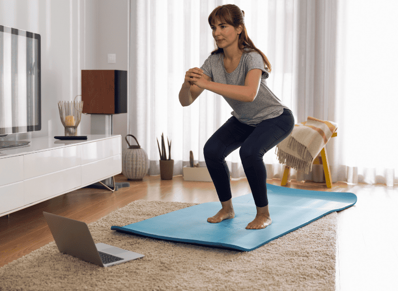 woman doing a squat on a yoga mat looking at a laptop on the floor