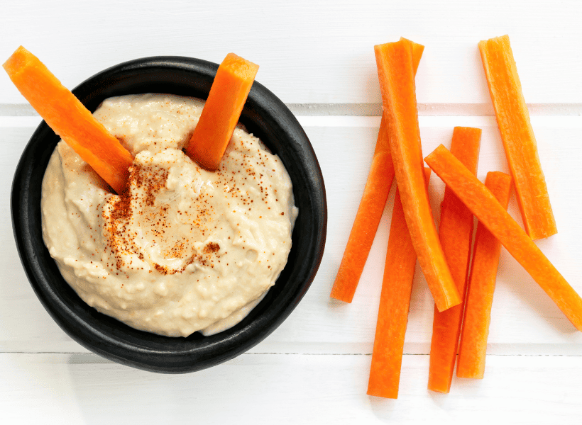 Overhead view of black bowl of hummus with carrots sticking in and carrots laying on table beside bowl.