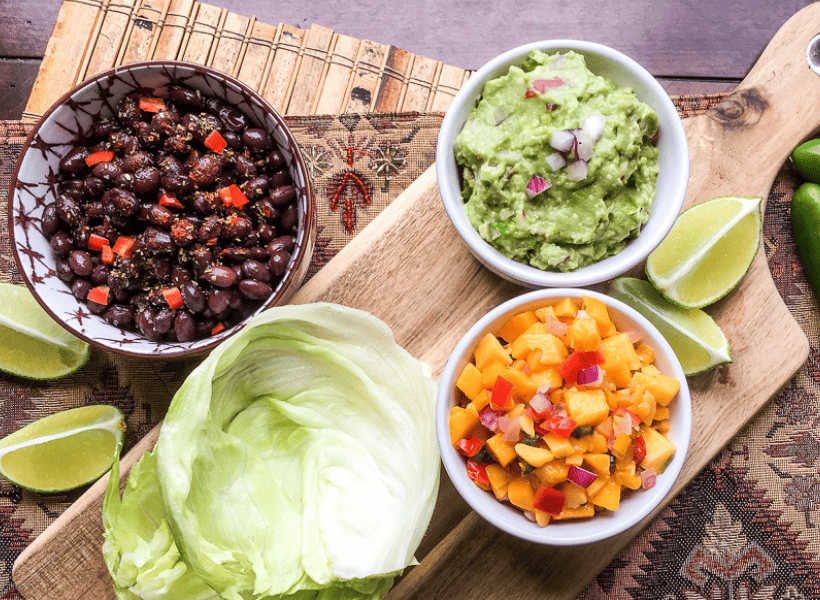 ingredients for a vegetarian lettuce wrap in white bowls on wooden cutting board mango salsa, guacamole, black beans and lettuce