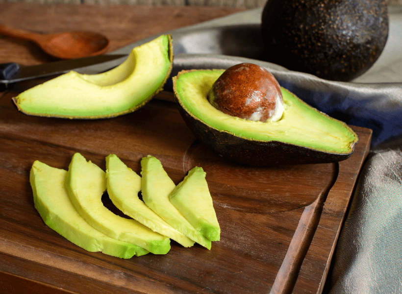avocado cut in half and sliced with another avocado beside it on a dark cutting board