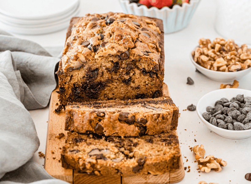 sliced chocolate chip zucchini bread on table beside bowls of chocolate chips and walnuts