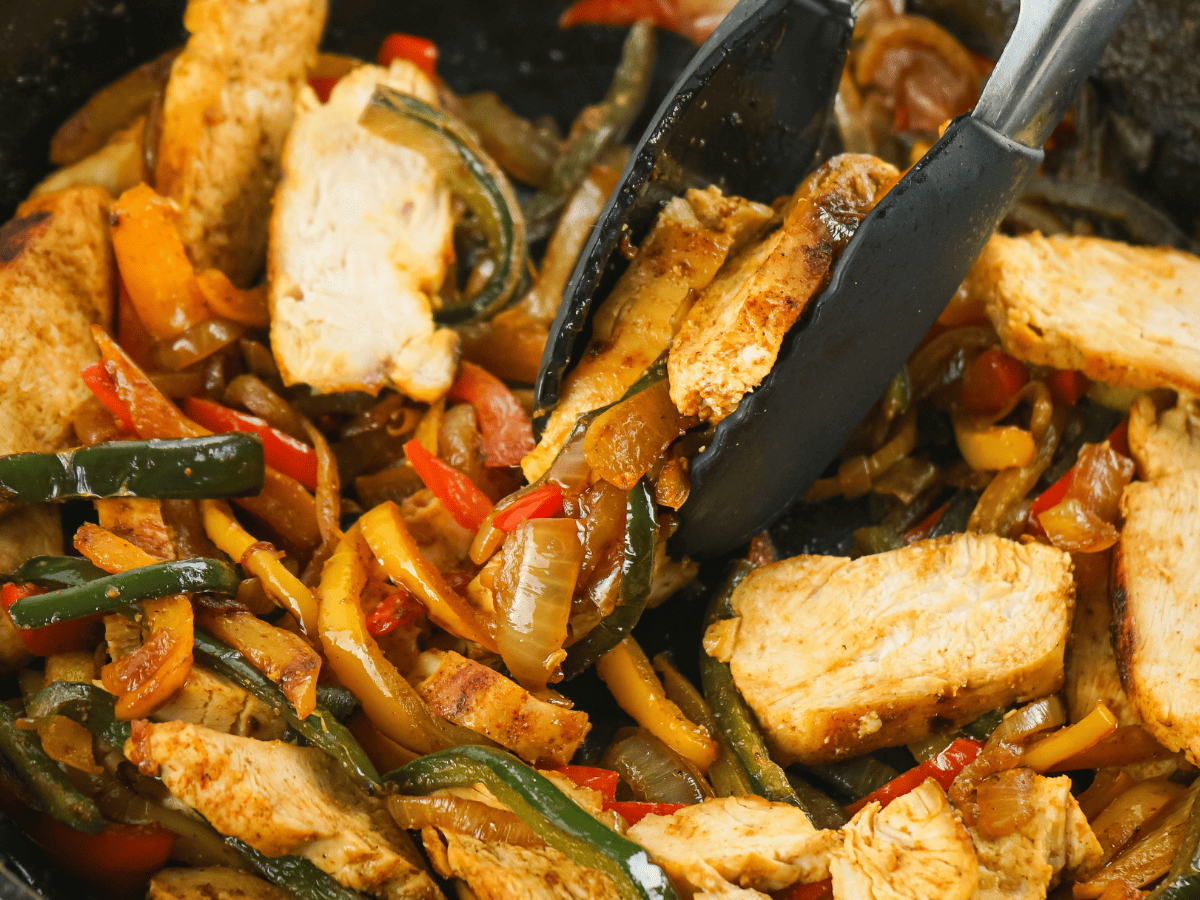 chicken fajitas cooked in cast iron skillet with tongs picking some up