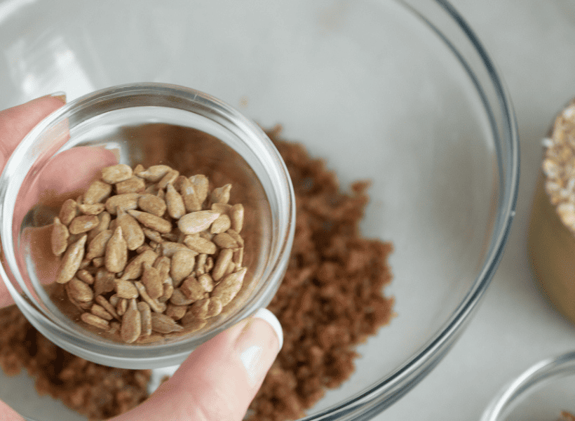 homemade granola mixture in glass bowl and sunflower seeds being added