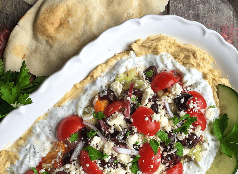 layered dip with hummus, olives, tomatoes, cheese, onions on a plate