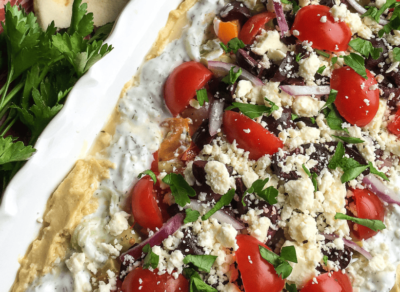 layered greek dip with olives, tomatoes, red onions, hummus on a plate