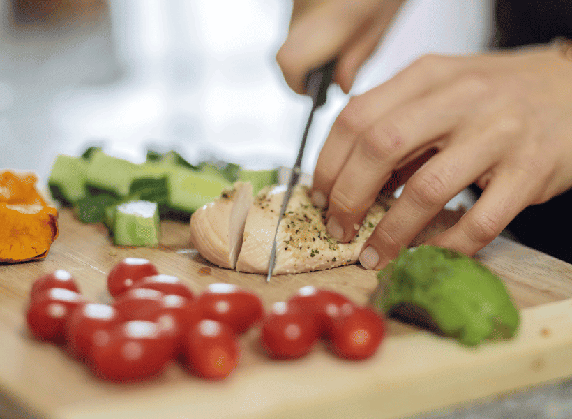 person chopping chicken beside chopped vegetables tomatoes cucumbers and peppers